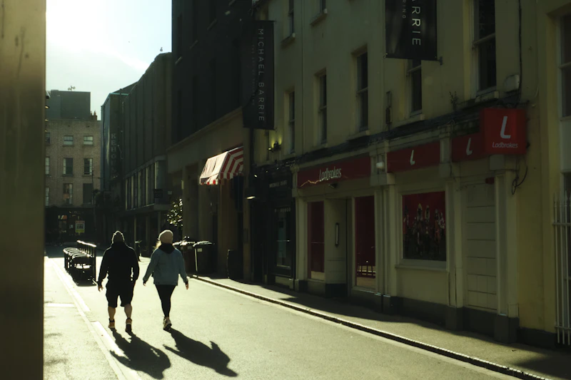 Two people walk down a sunny street with shadows.
