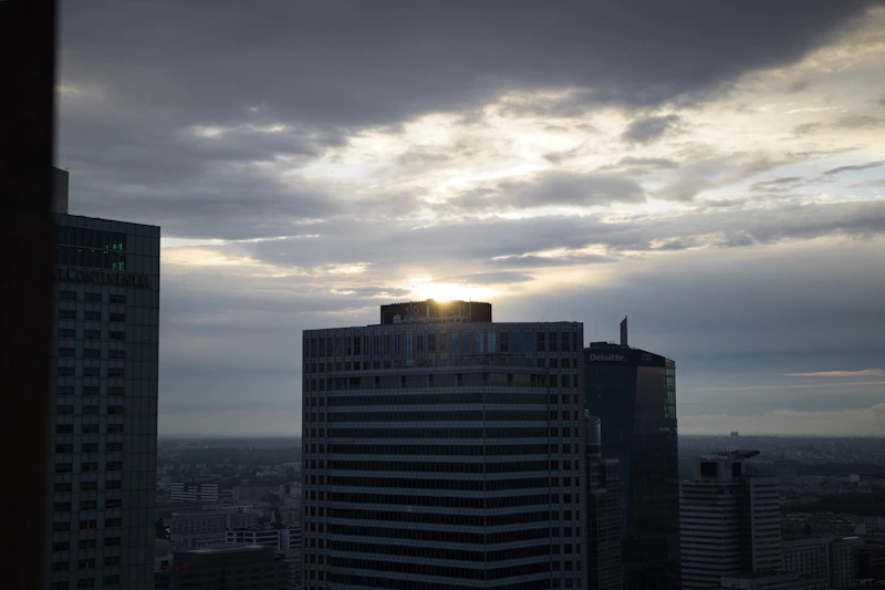 Sun setting behind modern city skyscrapers under cloudy sky.